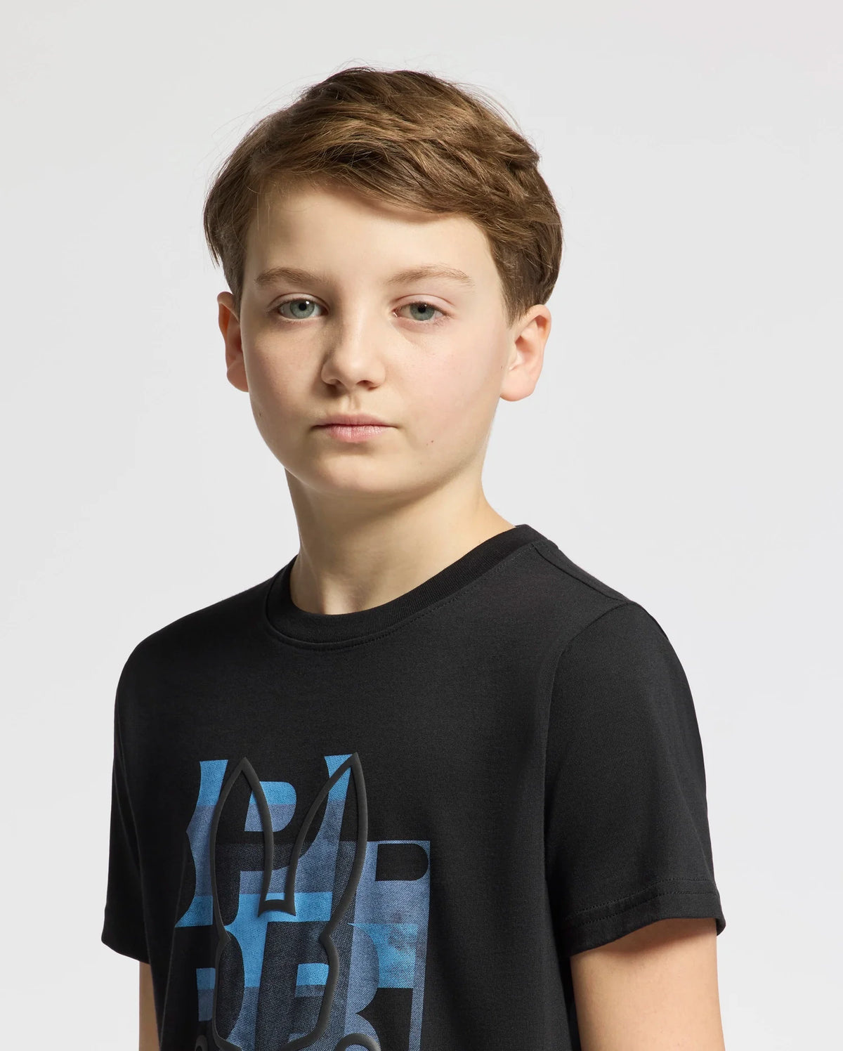 A young boy with short brown hair wears the black Psycho Bunny KIDS ZEALAND GRAPHIC TEE - B0U604F200, crafted from soft Pima cotton and featuring a blue abstract design with a subtle bunny logo. He stands against a white background, looking directly at the camera with a neutral expression.