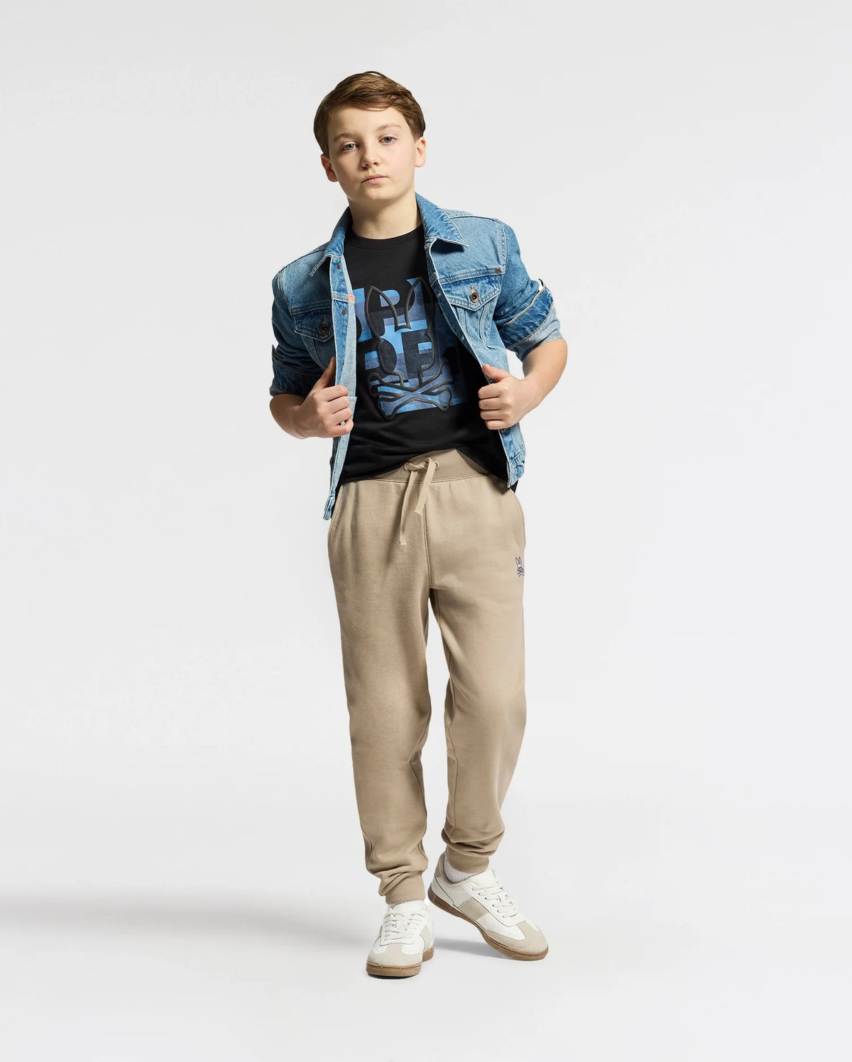 A young person stands wearing a denim jacket over the KIDS ZEALAND GRAPHIC TEE by Psycho Bunny, beige jogger pants, and white sneakers. They pose with hands in pockets against a plain white backdrop.
