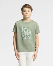 A boy with short light brown hair stands against a plain white background, wearing the KIDS ZEALAND GRAPHIC TEE - B0U604F200 by Psycho Bunny. This soft Pima cotton tee displays a big bunny logo with bones on the front and is paired with crisp white pants, matching his neutral expression.