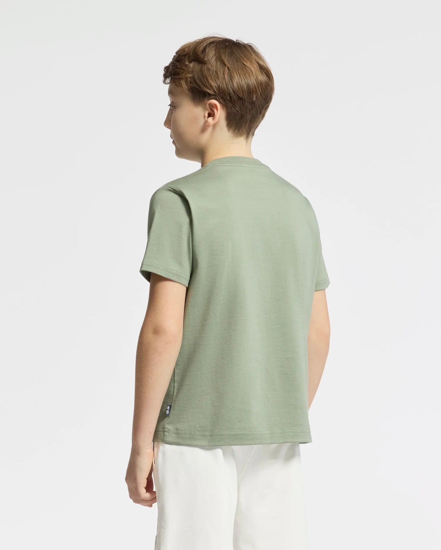 A boy is facing away from the camera, wearing a Psycho Bunny KIDS ZEALAND GRAPHIC TEE - B0U604F200 in light green with a large bunny logo and white shorts. He stands against a white background.