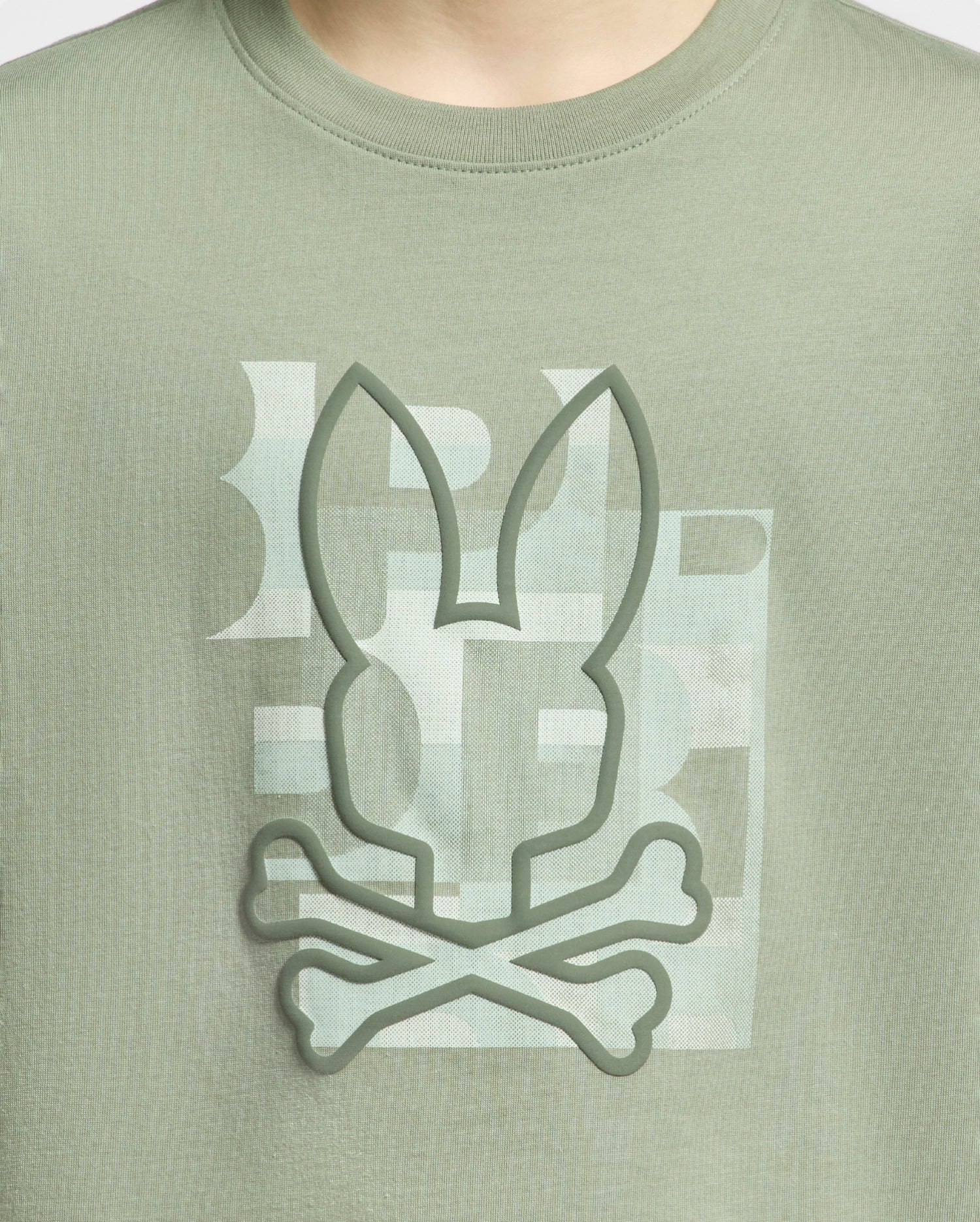 Close-up of a person wearing a Psycho Bunny KIDS ZEALAND GRAPHIC TEE - B0U604F200 in green Pima cotton, featuring a large raised bunny logo with crossbones design in a playful tone.
