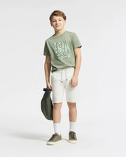 A boy in a Psycho Bunny KIDS ZEALAND GRAPHIC TEE - B0U604F200 stands against a white background. He's wearing white shorts, socks, and green sneakers, holding a matching green bag.