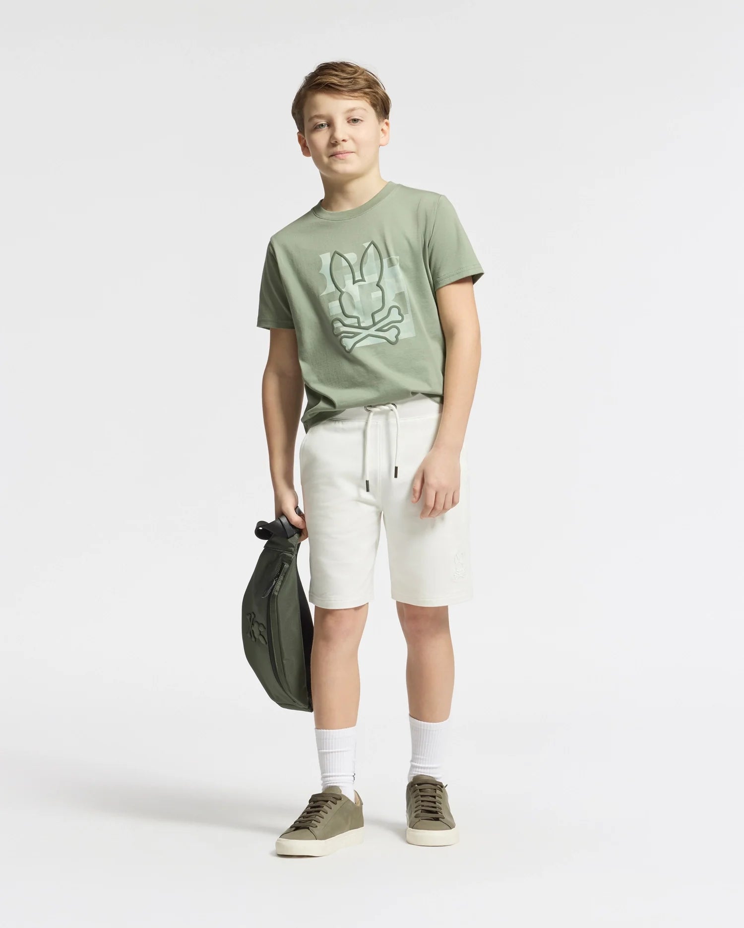 A boy in a Psycho Bunny KIDS ZEALAND GRAPHIC TEE - B0U604F200 stands against a white background. He's wearing white shorts, socks, and green sneakers, holding a matching green bag.