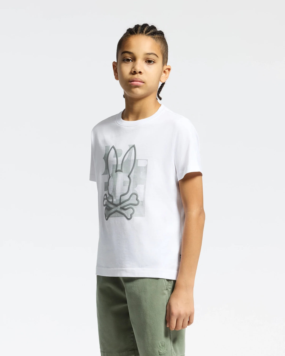 A young person with braided hair stands against a light background, wearing the white KIDS ZEALAND GRAPHIC TEE by Psycho Bunny. Crafted from soft Pima cotton, it features a big bunny skull logo and pairs perfectly with green pants.