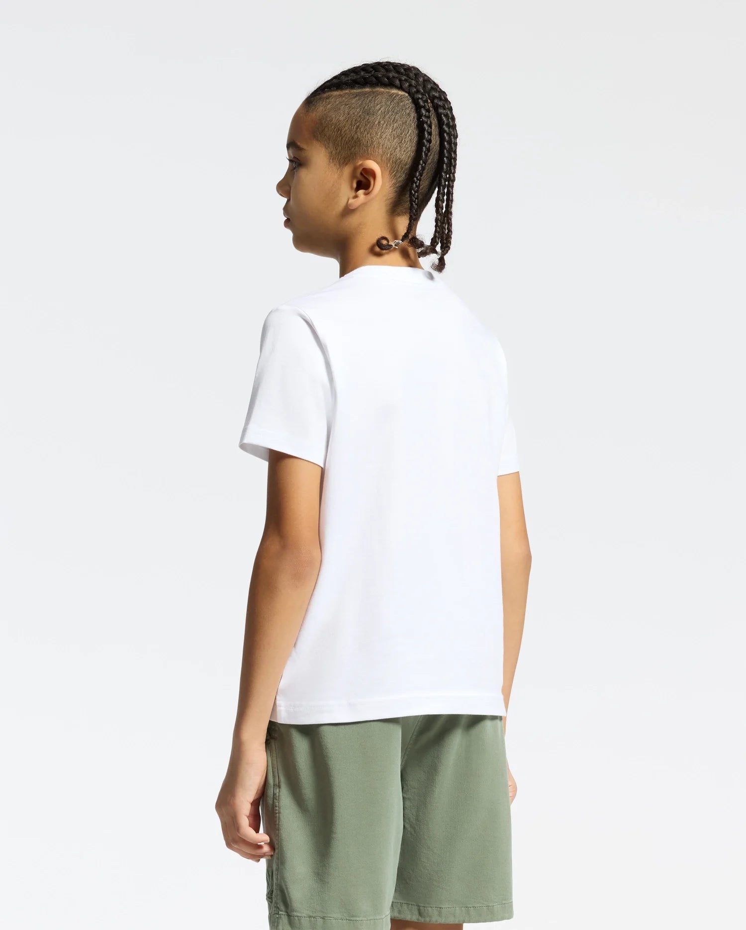 A person with braided hair faces away, wearing a Psycho Bunny KIDS ZEALAND GRAPHIC TEE in soft Pima cotton paired with green shorts. The background is plain white.