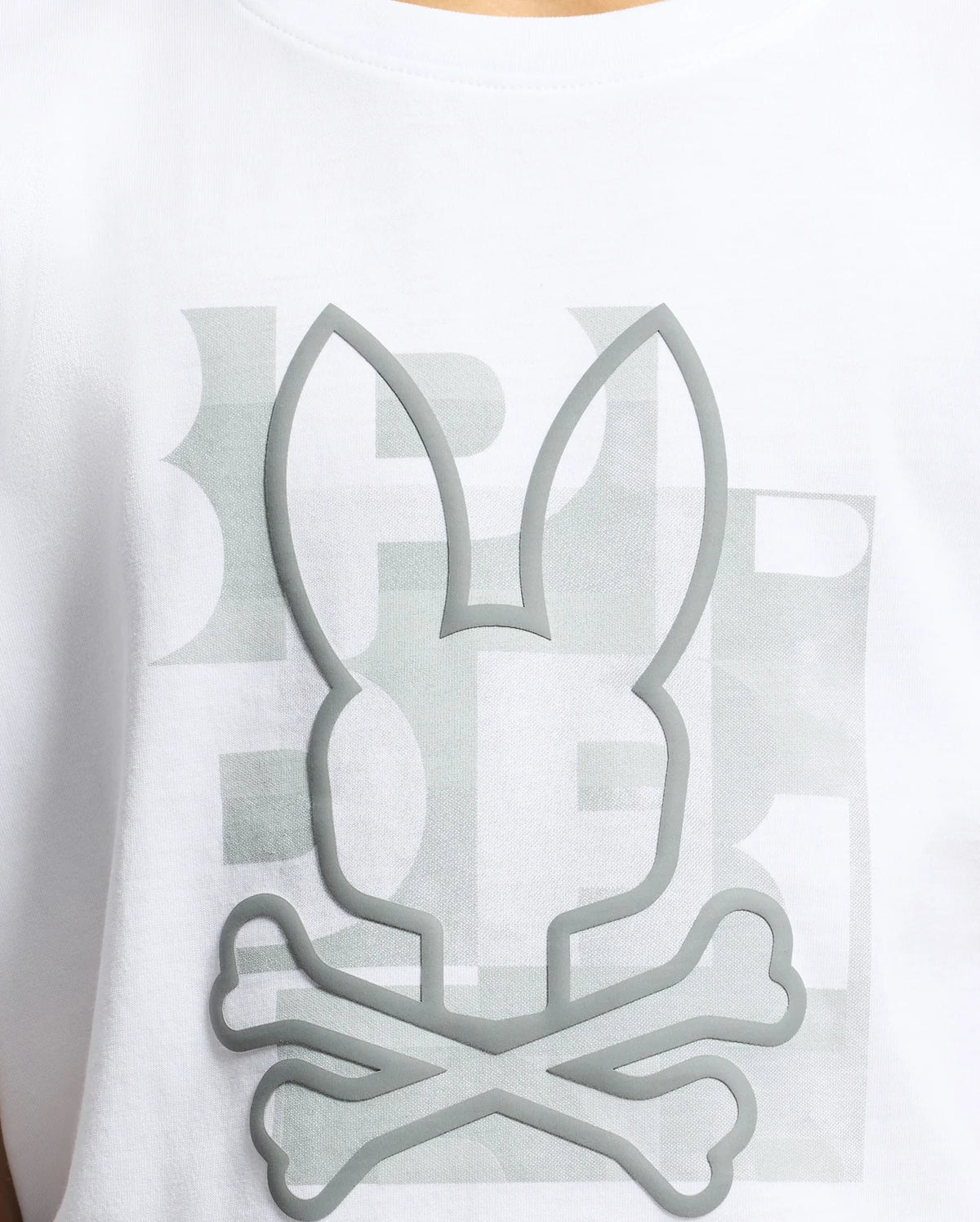Close-up of the KIDS ZEALAND GRAPHIC TEE by Psycho Bunny in soft Pima cotton, featuring a big bunny logo with crossbones below. The embossed gray design is positioned over faded background text.