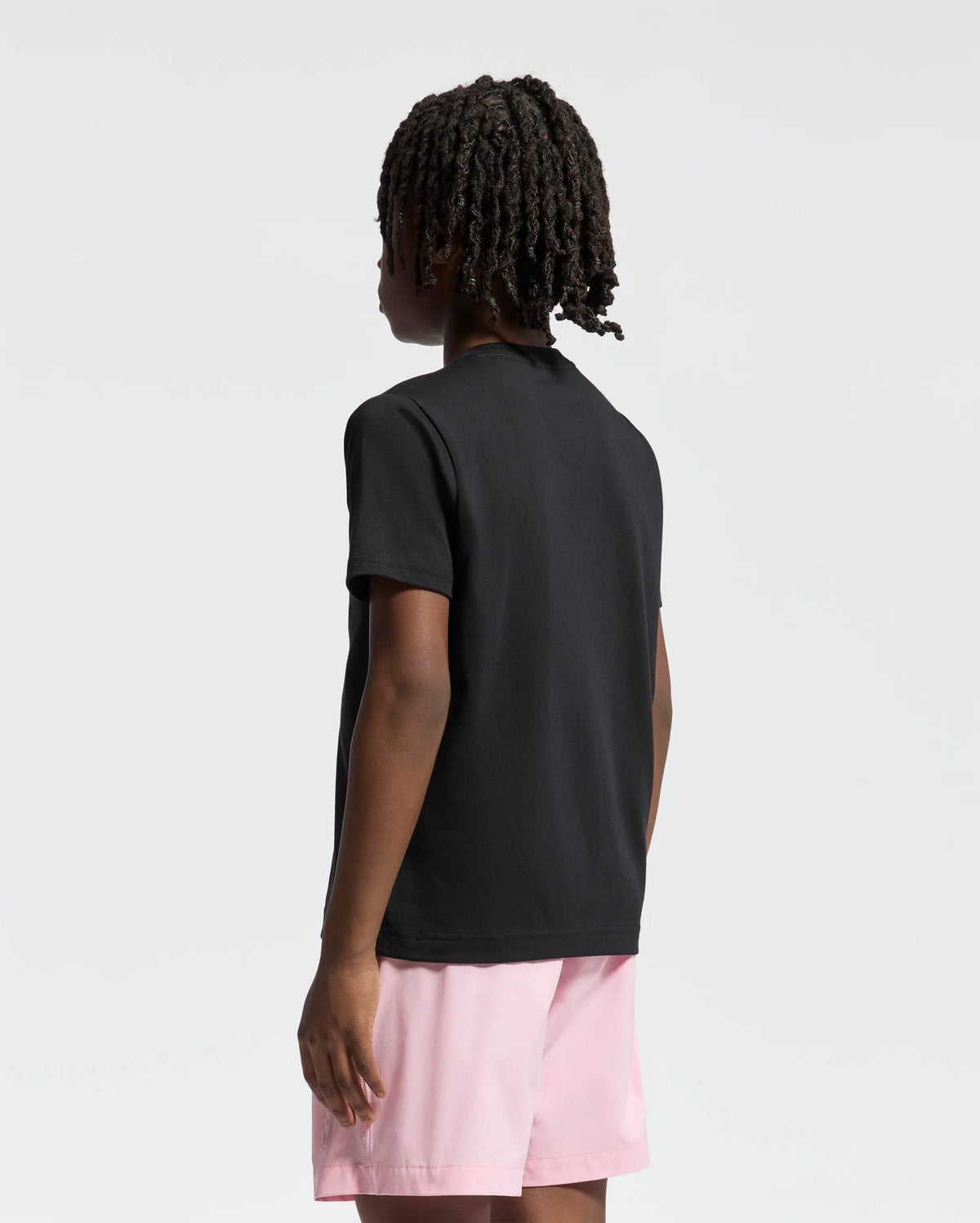A person with curly hair stands facing away, wearing a Psycho Bunny black tee featuring a bunny graphic and pink shorts against a plain, light-colored background.