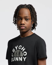 A person with short dreadlocks wears a black KIDS GOLDIE GRAPHIC TEE (B0U606F200) by Psycho Bunny, featuring a bunny graphic logo. Made from soft Peruvian Pima cotton, they have a neutral expression against a light gray background.