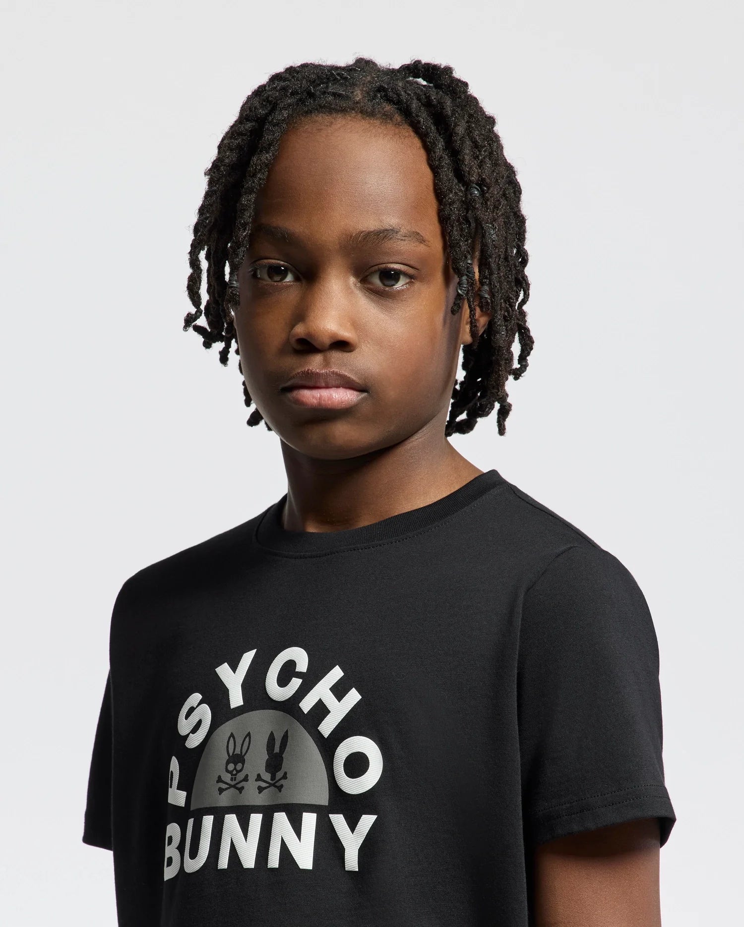 A person with short dreadlocks wears a black KIDS GOLDIE GRAPHIC TEE (B0U606F200) by Psycho Bunny, featuring a bunny graphic logo. Made from soft Peruvian Pima cotton, they have a neutral expression against a light gray background.
