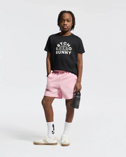 A child stands confidently against a plain background, wearing a comfortable KIDS GOLDIE GRAPHIC TEE from Psycho Bunny, light pink shorts, white socks with a dark symbol, and white sneakers. They hold a black water bottle in one hand.