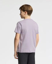 A young person with short brown hair, facing away, wears a light purple KIDS GOLDIE GRAPHIC TEE by Psycho Bunny. Made from soft Peruvian Pima cotton and featuring a subtle bunny graphic, the tee complements their black pants against the plain white background.