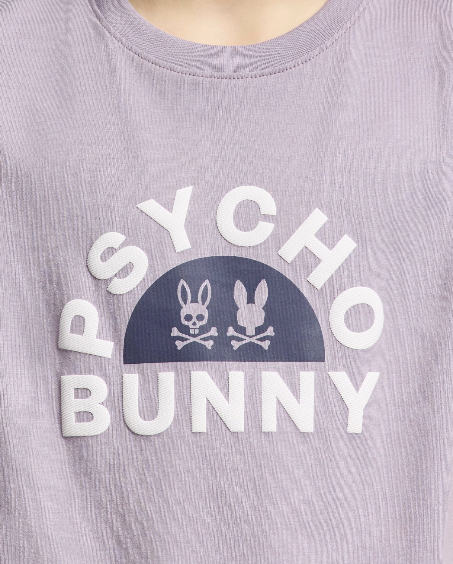 A close-up of the KIDS GOLDIE GRAPHIC TEE by Psycho Bunny, made from luxurious purple Peruvian Pima cotton, features bold white "PSYCHO BUNNY" text with unique stylized bunny graphics and crossed bones.