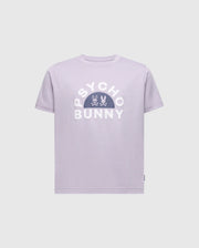 The KIDS GOLDIE GRAPHIC TEE by Psycho Bunny is made of soft Peruvian Pima cotton. The design showcases "PSYCHO BUNNY" in bold white letters, with two small bunny skull icons centered in a semicircle below, all on a lavender background.