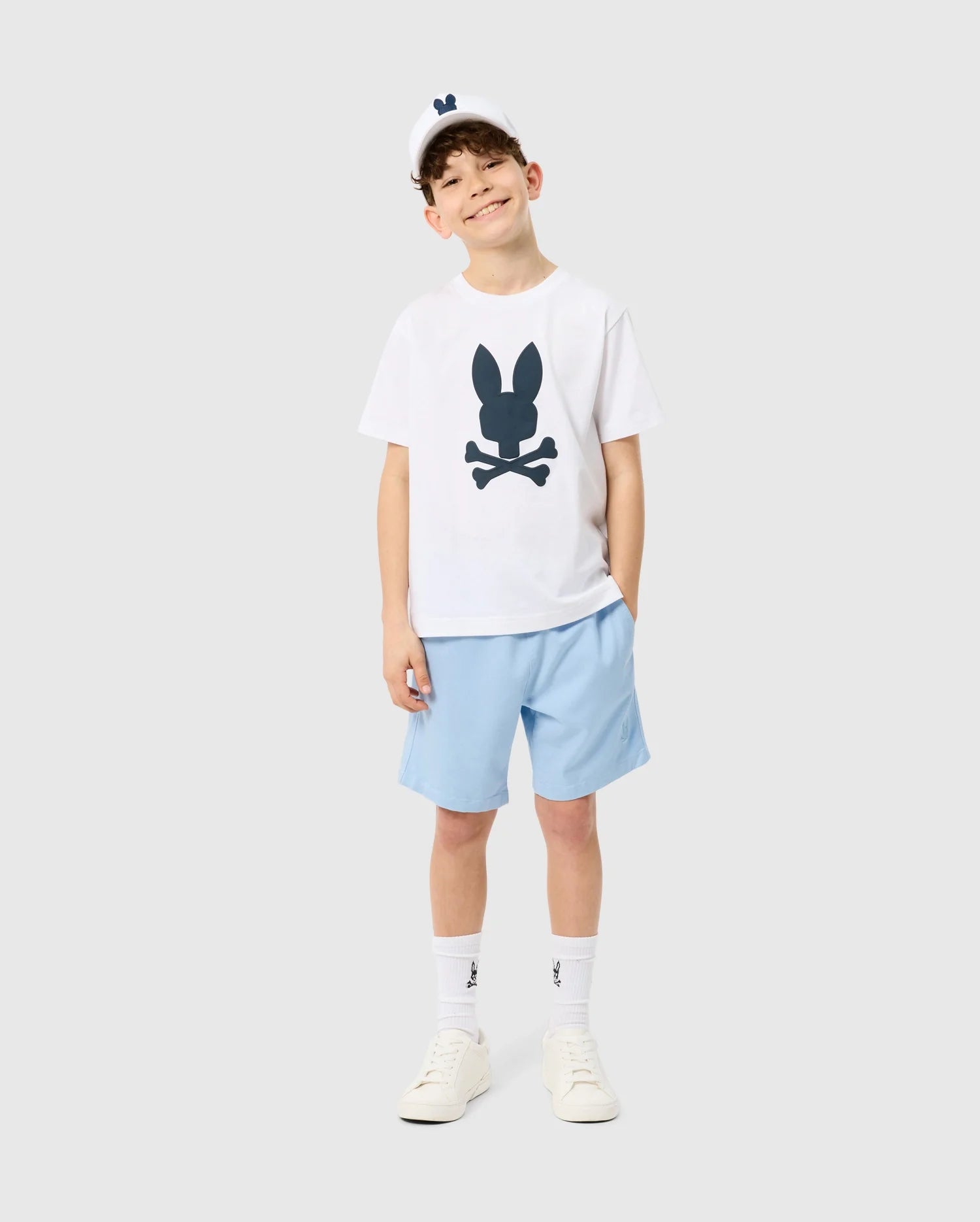 A young boy posing with a smile, wearing a Psycho Bunny KIDS HOUSTON GRAPHIC TEE - B0U607C200, light blue shorts, white socks, and sneakers, standing against a plain light background.