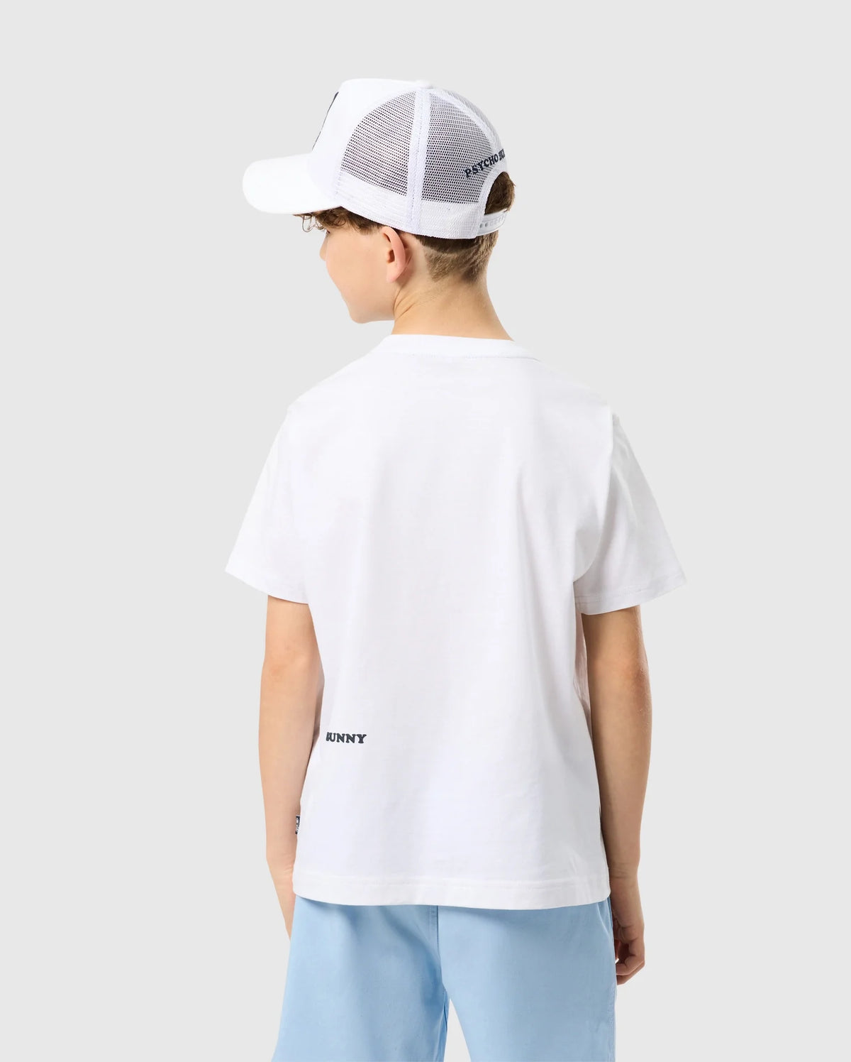 A young person from behind wearing a Psycho Bunny Kids Houston Graphic Tee and a white mesh-back cap, standing against a light gray background.