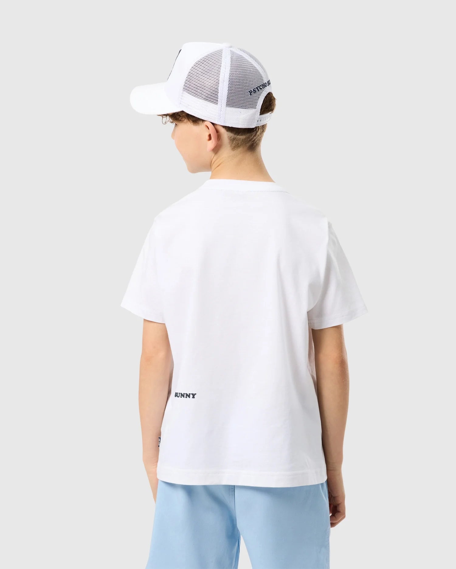 A young person from behind wearing a Psycho Bunny Kids Houston Graphic Tee and a white mesh-back cap, standing against a light gray background.
