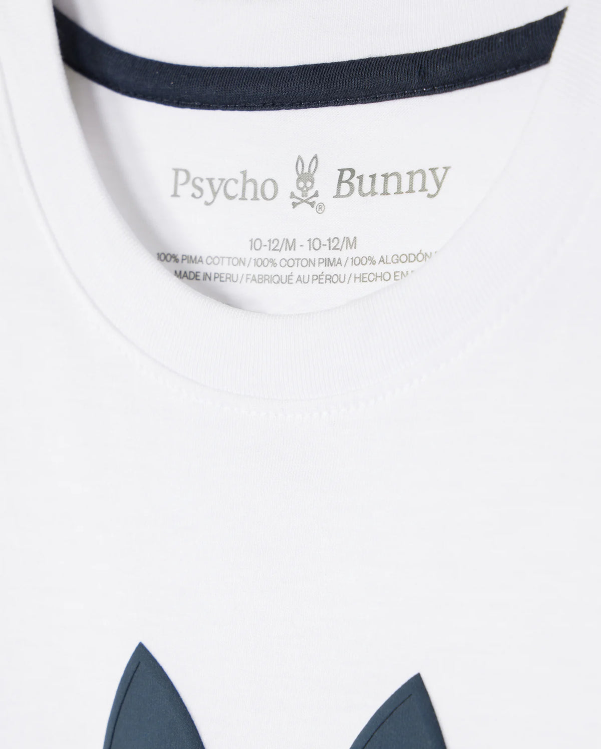 Close-up of a white KIDS HOUSTON GRAPHIC TEE - B0U607C200 tag with the brand "Psycho Bunny" printed above wash instructions and fabric composition, with a hint of the garment's navy detail visible.