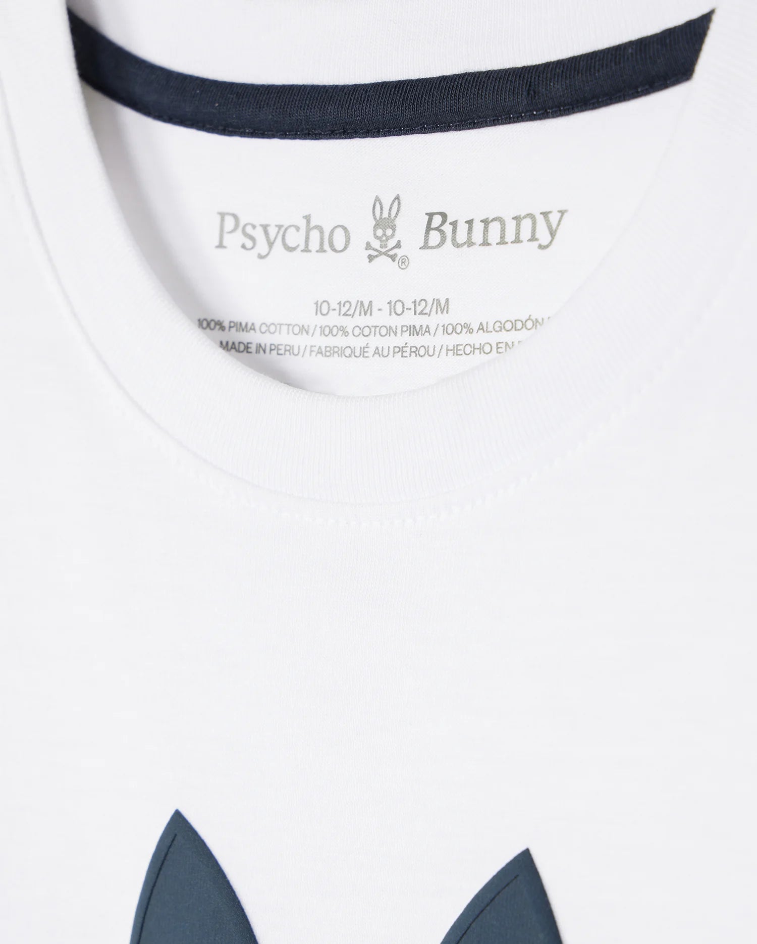 Close-up of a white KIDS HOUSTON GRAPHIC TEE - B0U607C200 tag with the brand "Psycho Bunny" printed above wash instructions and fabric composition, with a hint of the garment's navy detail visible.