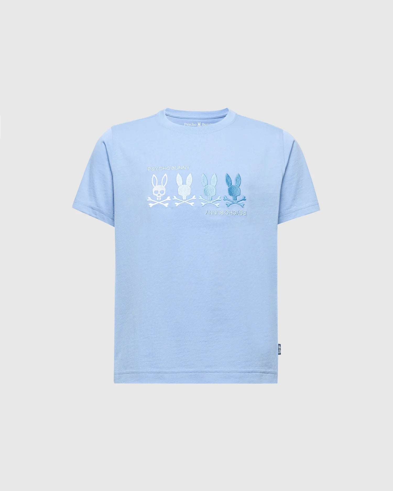 The KIDS OCEANIA EMBROIDERED GRAPHIC TEE by Psycho Bunny, product B0U607F200, features retro light blue Pima cotton with embroidered bunny skull and crossbones in varying shades. Blue bunnies are on the right and a white bunny on the left. A small label decorates the bottom left hem.