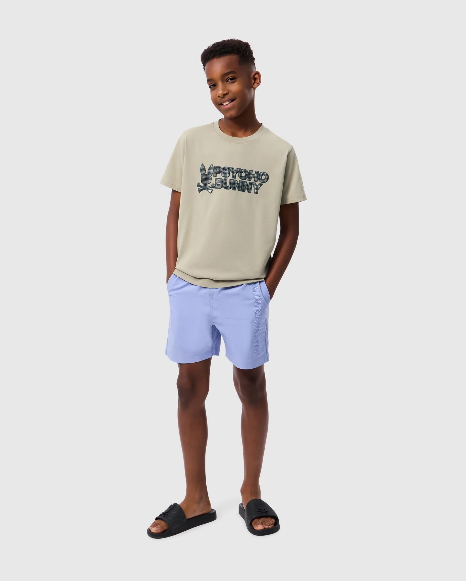 A young person stands with hands in pockets, wearing a KIDS MELVIN GRAPHIC TEE by Psycho Bunny, made from Pima cotton and featuring a wordmark graphic print. They are also dressed in light blue shorts and black slide sandals. The individual has short hair and the background is plain white.