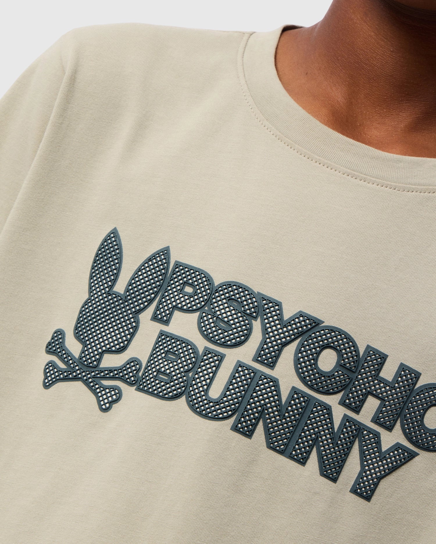Close-up of a child wearing the KIDS MELVIN GRAPHIC TEE by Psycho Bunny, in beige Pima cotton. The t-shirt showcases a stylized graphic design of a bunny head with crossbones and the text "PSYCHO BUNNY" in textured dark print. The wordmark graphic is rendered in an HD-silicone print on a plain background, focusing on the chest area.