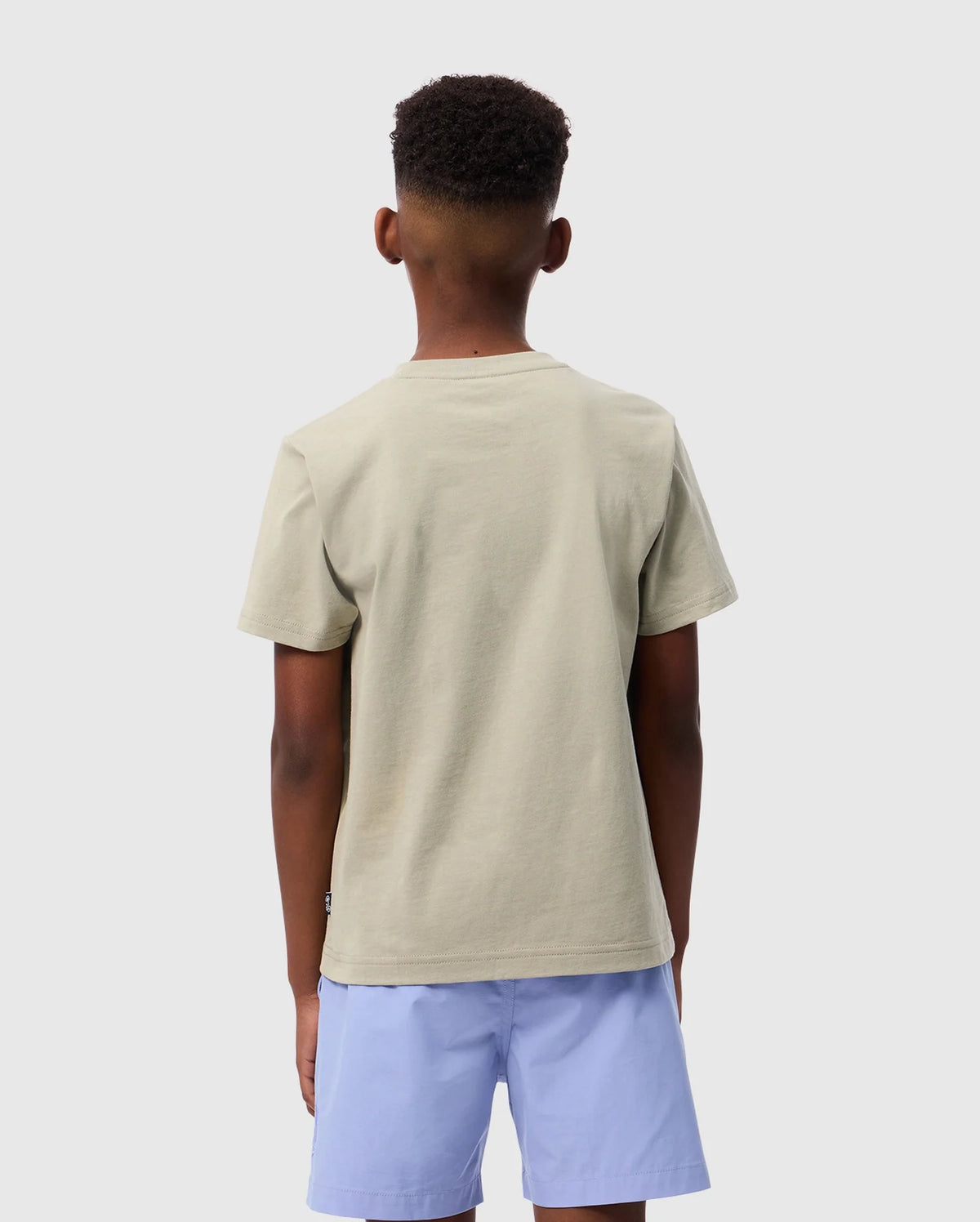 A young person with short hair is facing away from the camera, wearing a light beige Psycho Bunny KIDS MELVIN GRAPHIC TEE - B0U663C200 featuring a subtle HD-silicone print and light blue shorts. The background is plain white.