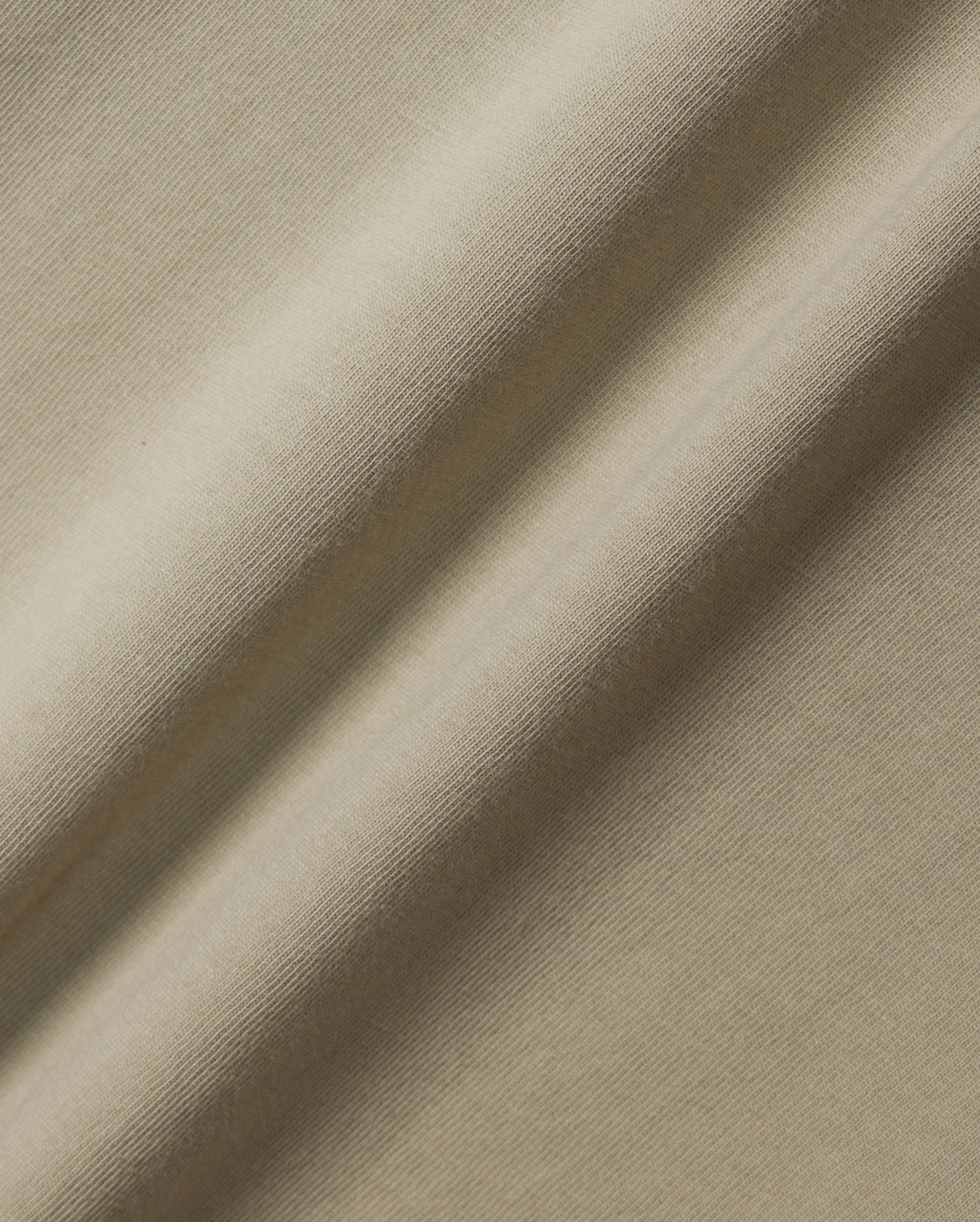 A close-up of the beige Pima cotton fabric of the KIDS MELVIN GRAPHIC TEE - B0U663C200 from Psycho Bunny, showcasing its smooth texture and subtle diagonal folds. The lighting highlights the gentle sheen and soft appearance of this textile.