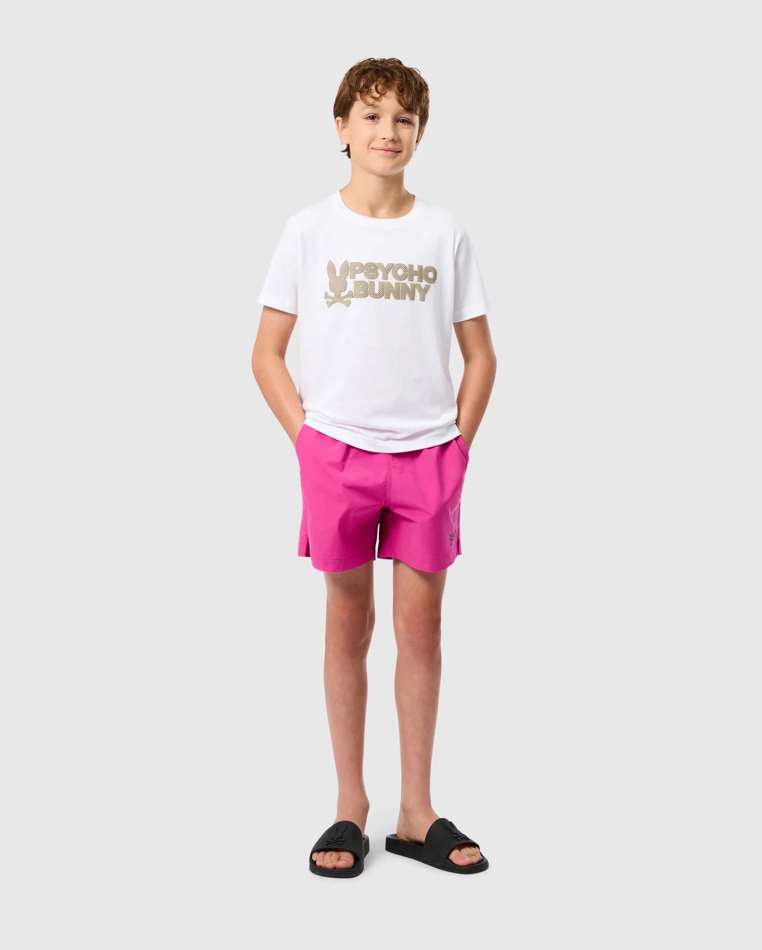 A young person with short brown hair stands against a plain background. They are wearing the KIDS MELVIN GRAPHIC TEE by Psycho Bunny, a white Pima cotton t-shirt featuring a graphic and text that reads "Psycho Bunny," paired with bright pink shorts. They also wear black slide sandals and have their hands in their pockets.