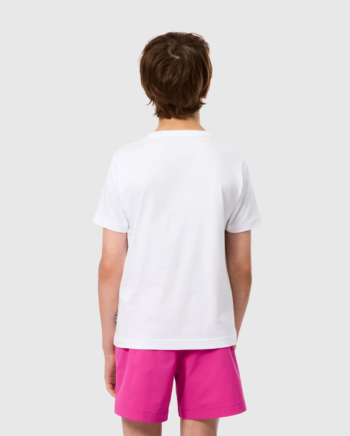 A person with short brown hair is seen from the back, wearing a KIDS MELVIN GRAPHIC TEE by Psycho Bunny and bright pink shorts. The background is a solid light gray.