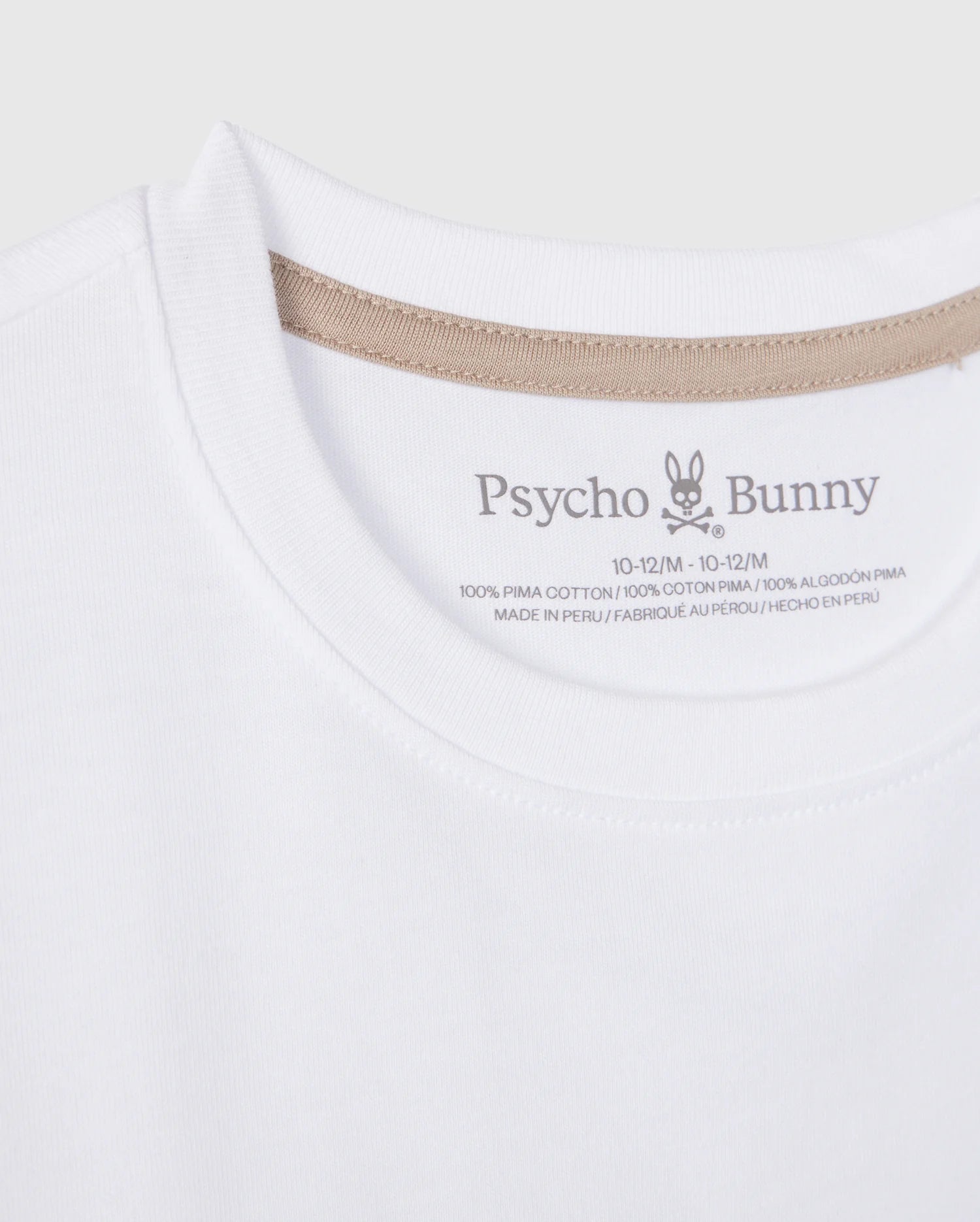 A close-up photo of a white Psycho Bunny KIDS MELVIN GRAPHIC TEE - B0U663C200. The image focuses on the collar area, showing brand details and size information, with a Bunny with wordmark printed on the inner neck label. The shirt is made of 100% Pima cotton and it is made in Peru.