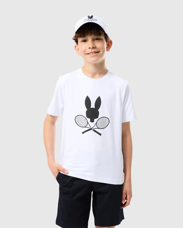 A smiling boy is standing against a plain light gray background. He wears a Psycho Bunny KIDS COURTSIDE GRAPHIC TEE - B0U681C200 featuring a black bunny and crossed tennis racket design, black shorts, and a white baseball cap. With his left hand in his pocket, he shows off his tennis-inspired look, complete with short, curly brown hair.