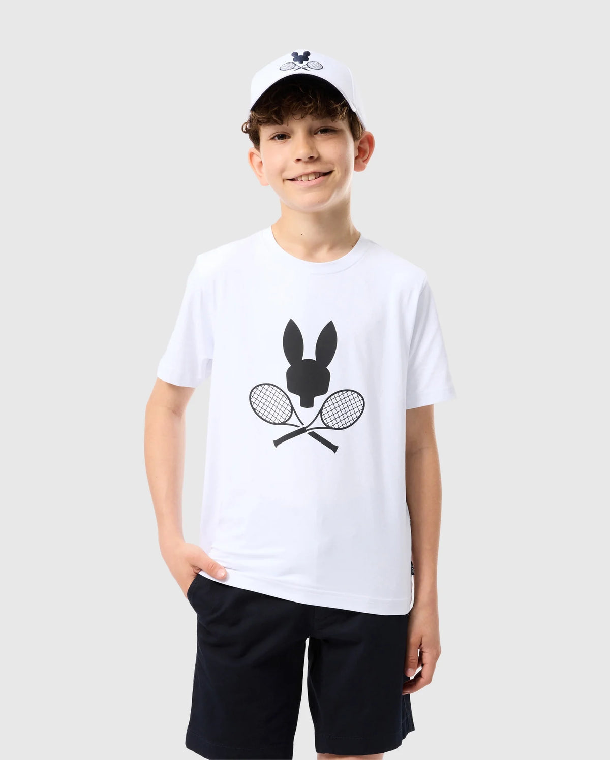 A smiling boy is standing against a plain light gray background. He wears a Psycho Bunny KIDS COURTSIDE GRAPHIC TEE - B0U681C200 featuring a black bunny and crossed tennis racket design, black shorts, and a white baseball cap. With his left hand in his pocket, he shows off his tennis-inspired look, complete with short, curly brown hair.