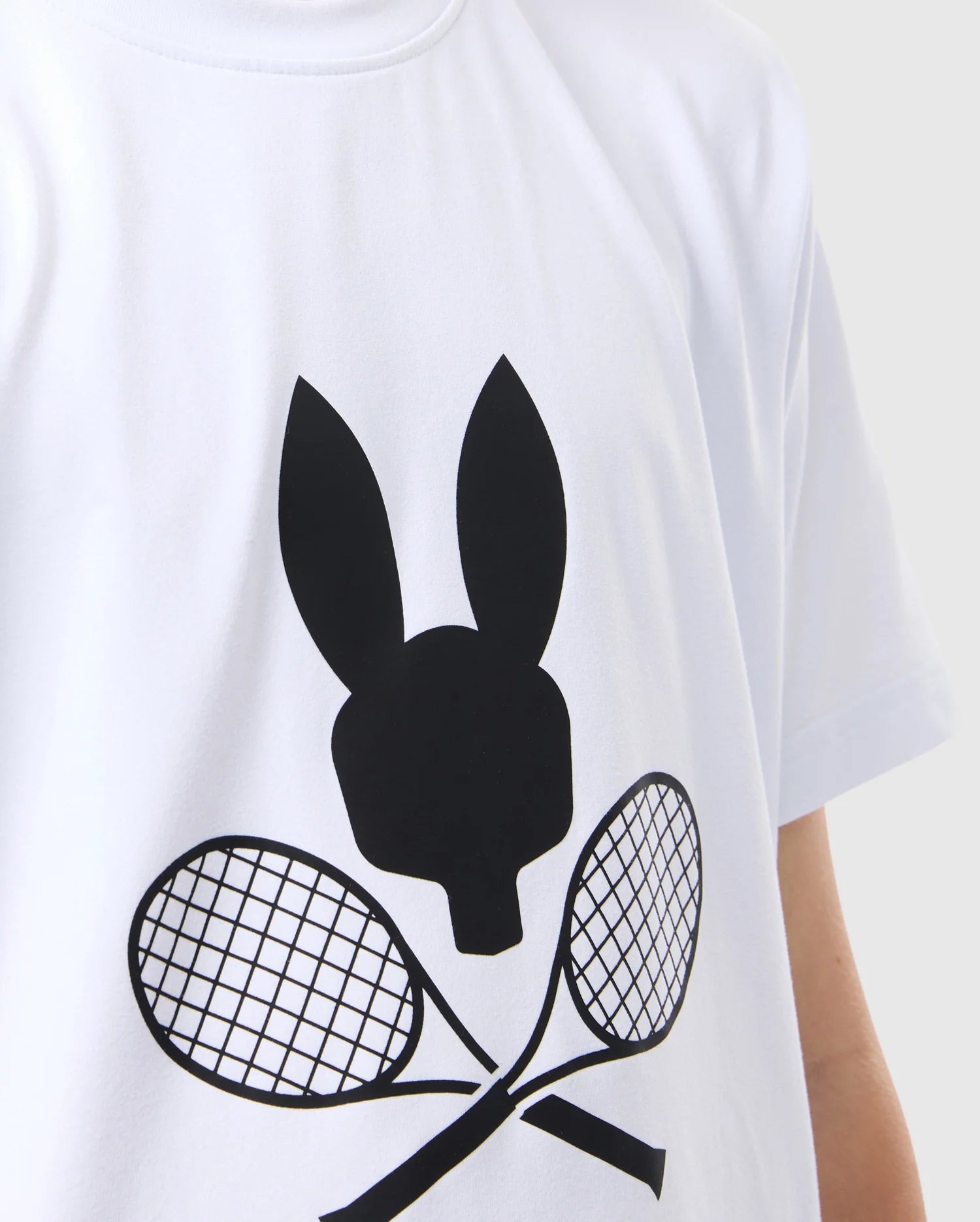 Close-up of a person wearing a white, tennis-inspired KIDS COURTSIDE GRAPHIC TEE - B0U681C200 from Psycho Bunny featuring a black graphic design. The graphic depicts a rabbit head silhouette above two crossed tennis rackets. The photo is cropped from the shoulder to the upper waist, showcasing this performance-led jersey.
