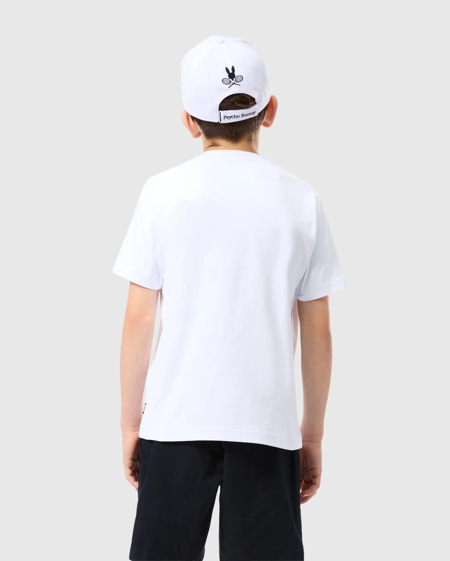 A young boy is standing with his back to the camera. He is wearing a white, KIDS COURTSIDE GRAPHIC TEE - B0U681C200 by Psycho Bunny, black shorts, and a white cap with a black graphic on the back. The background is plain, light grey.
