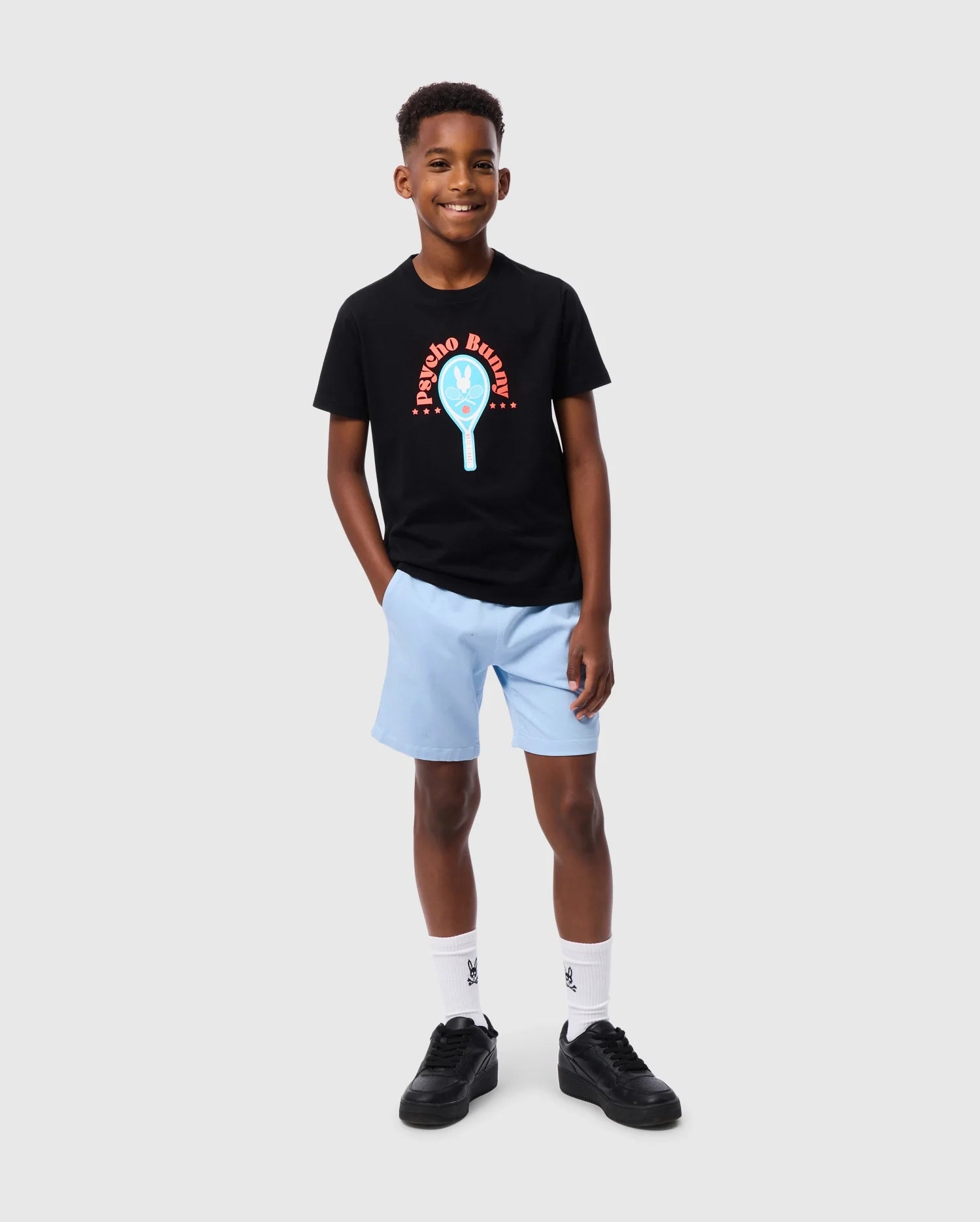 A young boy stands smiling, dressed in the Psycho Bunny KIDS COURTSIDE GRAPHIC TEE - B0U682C200, which showcases a vibrant design and text. He pairs it with light blue shorts, white socks, and black shoes. With his left hand casually tucked in his pocket against a plain gray background, his ensemble exudes an effortlessly tennis-inspired vibe.