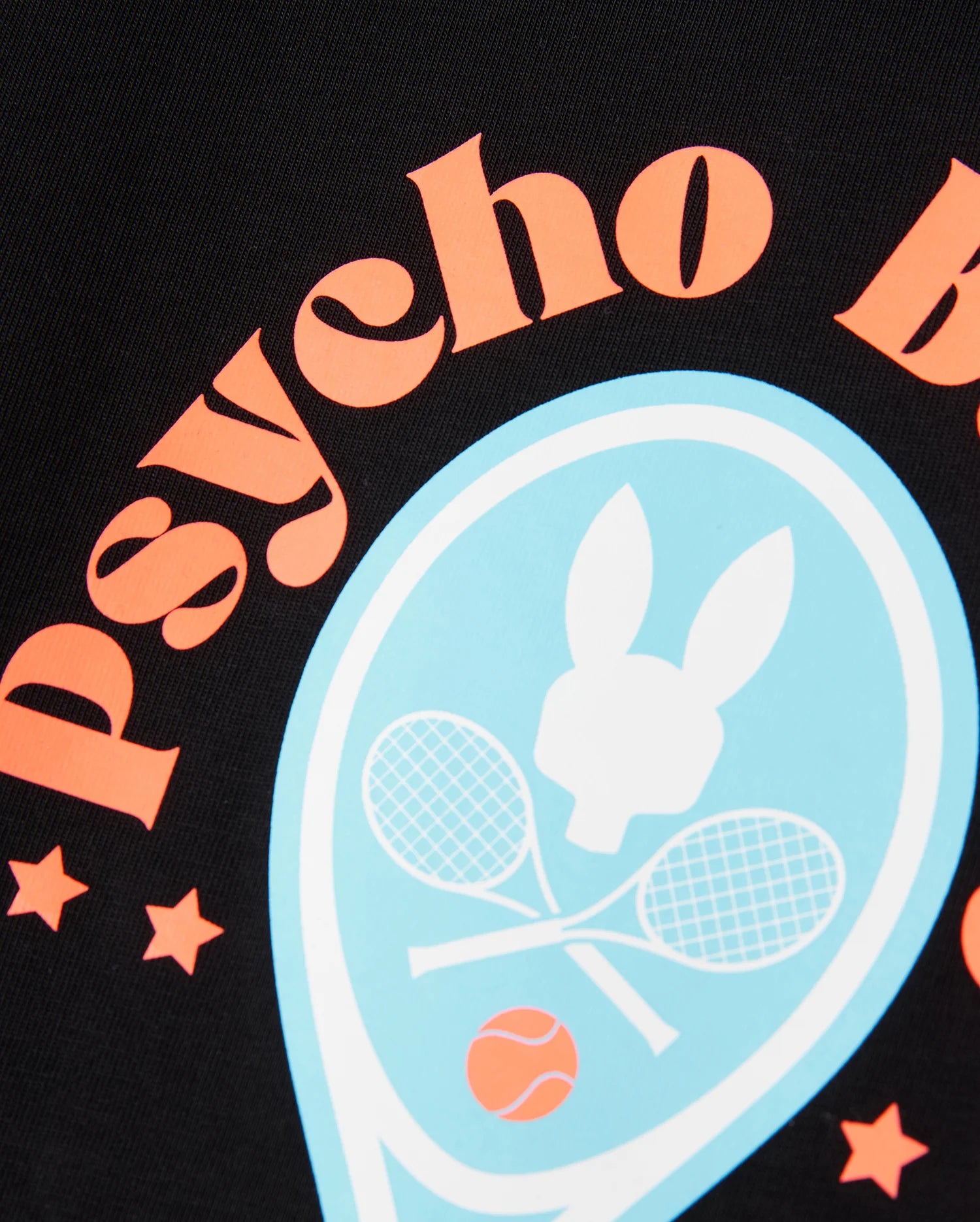 A close-up of a black fabric showcases the text "psycho b" in orange, arranged in an arc. At the center, there's a light blue emblem with a silhouette of a bunny's head above two crossed tennis rackets and a tennis ball. This performance-focused jersey by Psycho Bunny is bordered with orange stars, making the KIDS COURTSIDE GRAPHIC TEE - B0U682C200 perfect for kids' tennis-inspired adventures.