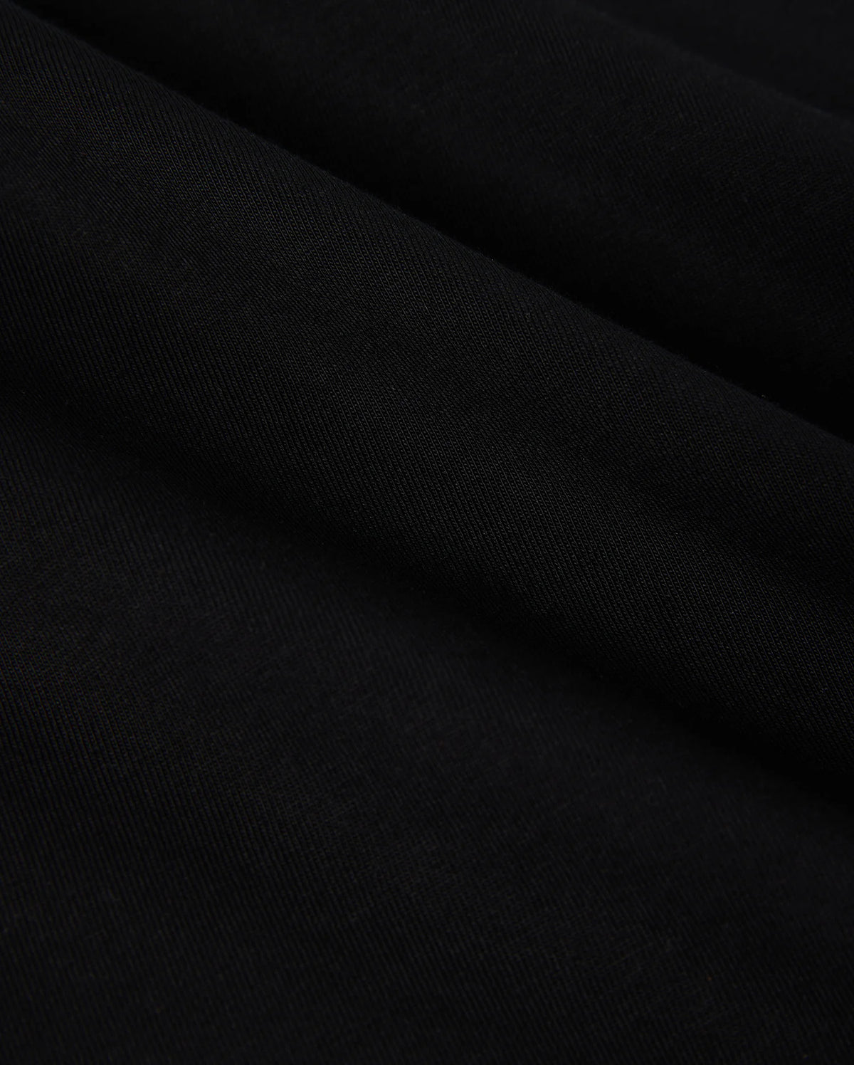 Close-up of the smooth, folded fabric of the KIDS COURTSIDE GRAPHIC TEE by Psycho Bunny in black, showcasing a subtle texture akin to a performance-led jersey. The material looks soft and slightly glossy, with gentle curves creating lines across the surface.