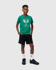 A young boy stands smiling against a plain white background. He is wearing a green Psycho Bunny KIDS COURTSIDE GRAPHIC TEE - B0U684C200 with a tennis club graphic, black shorts, white socks, and black sneakers. His hands are casually placed in his t-shirt pockets.