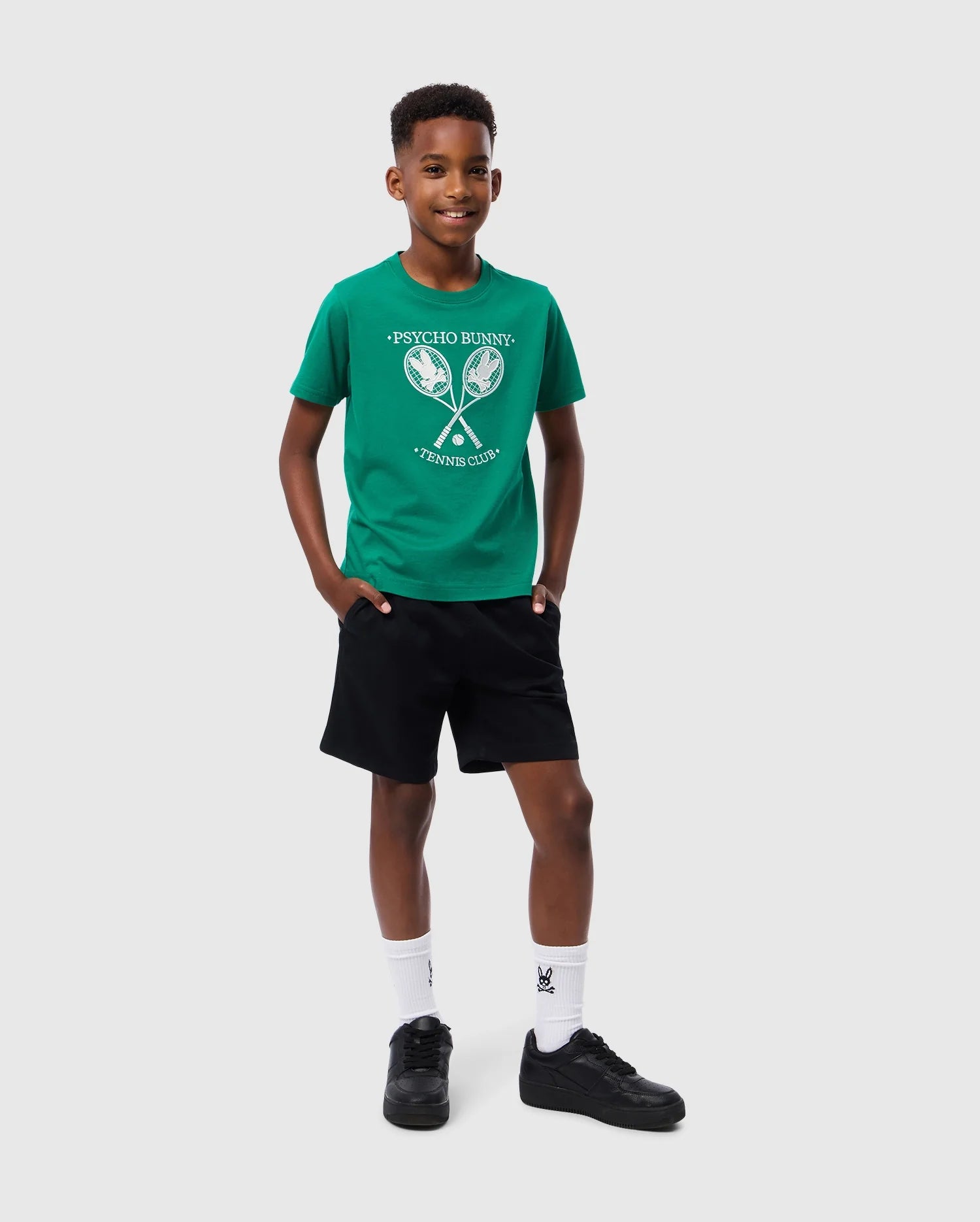 A young boy stands smiling against a plain white background. He is wearing a green Psycho Bunny KIDS COURTSIDE GRAPHIC TEE - B0U684C200 with a tennis club graphic, black shorts, white socks, and black sneakers. His hands are casually placed in his t-shirt pockets.
