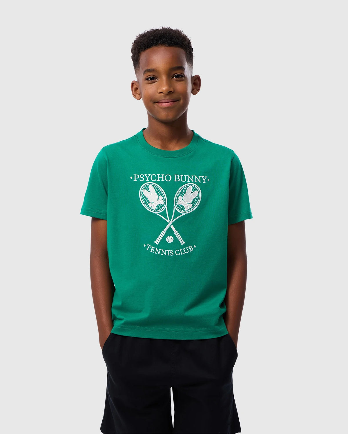 A young boy stands against a plain background wearing the Psycho Bunny KIDS COURTSIDE GRAPHIC TEE - B0U684C200 in green, featuring a "Psycho Bunny Tennis Club" print with two crossed tennis rackets. He has short, curly hair and dark shorts. He is smiling with his hands in his pockets, showcasing the stylish Pima cotton jersey.