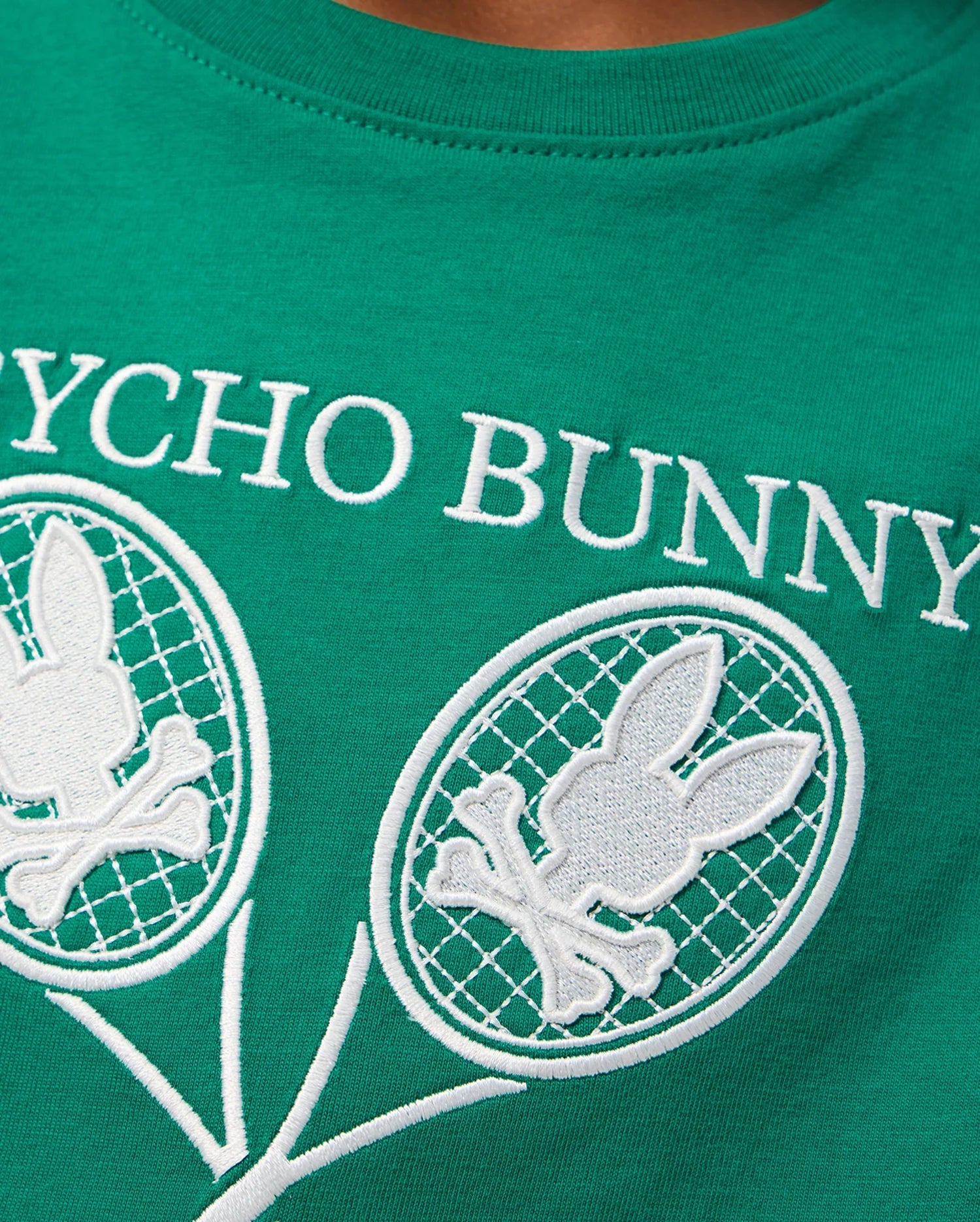 A close-up of a green Pima cotton jersey t-shirt from Psycho Bunny, featuring the text "Psycho Bunny" in white letters. Below the text are two white graphic designs of tennis rackets with bunny skull and crossbones symbols inside the racket heads, perfect for a Kids Courtside Graphic Tee - B0U684C200.
