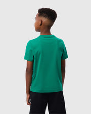 A young person with short hair is seen from behind, wearing a bright green Psycho Bunny Kids Courtside Graphic Tee (B0U684C200) and black shorts. The Pima cotton jersey material of the T-shirt offers a soft finish. They are standing against a plain light grey background.