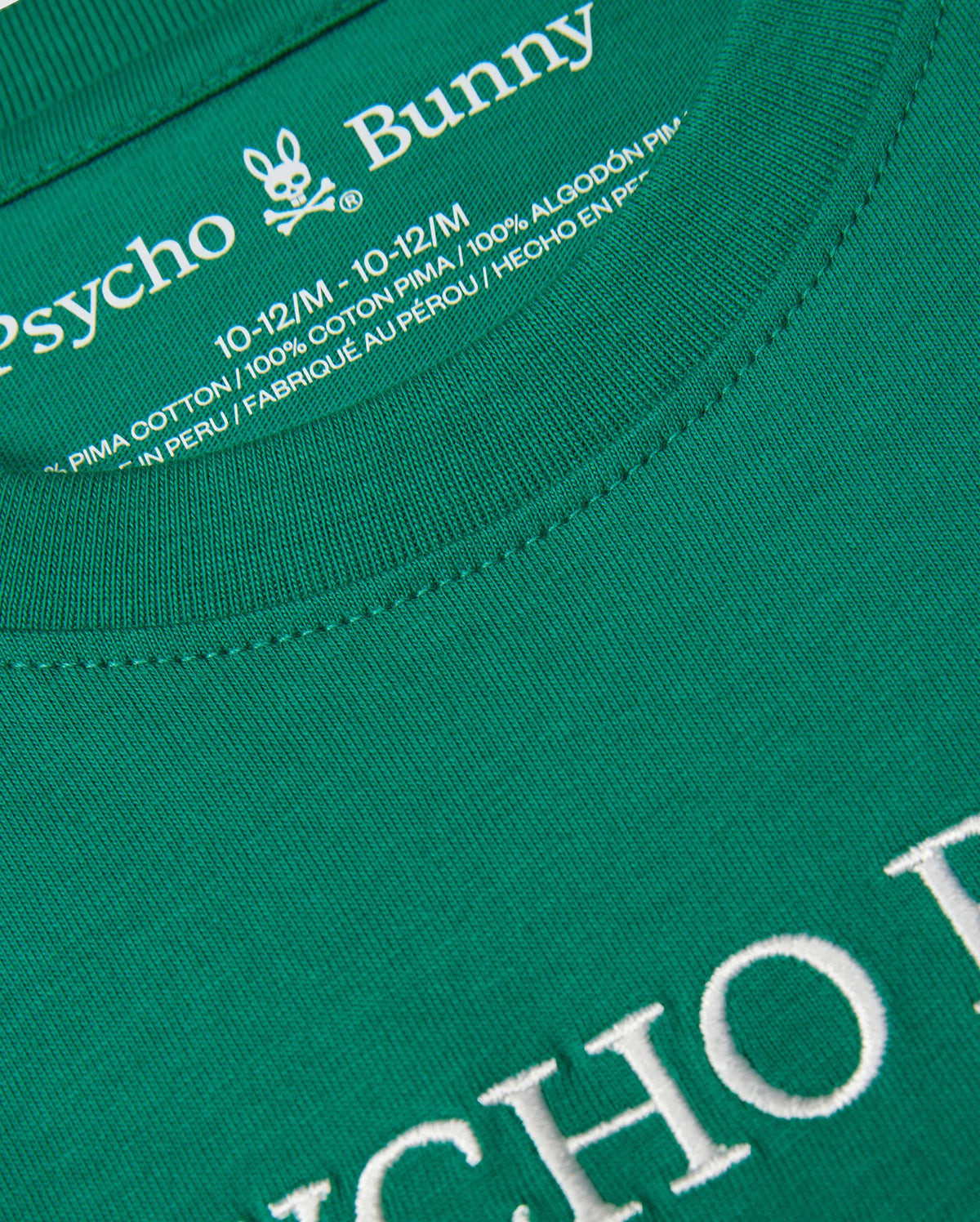 A close-up of a green KIDS COURTSIDE GRAPHIC TEE - B0U684C200 by Psycho Bunny. The text on the shirt's collar reads "Psycho Bunny" with a bunny logo above it. Below it states, "10-12/M, 100% Pima cotton jersey, Made in Peru." The shirt has white embroidered text that is partially visible at the bottom of the image.