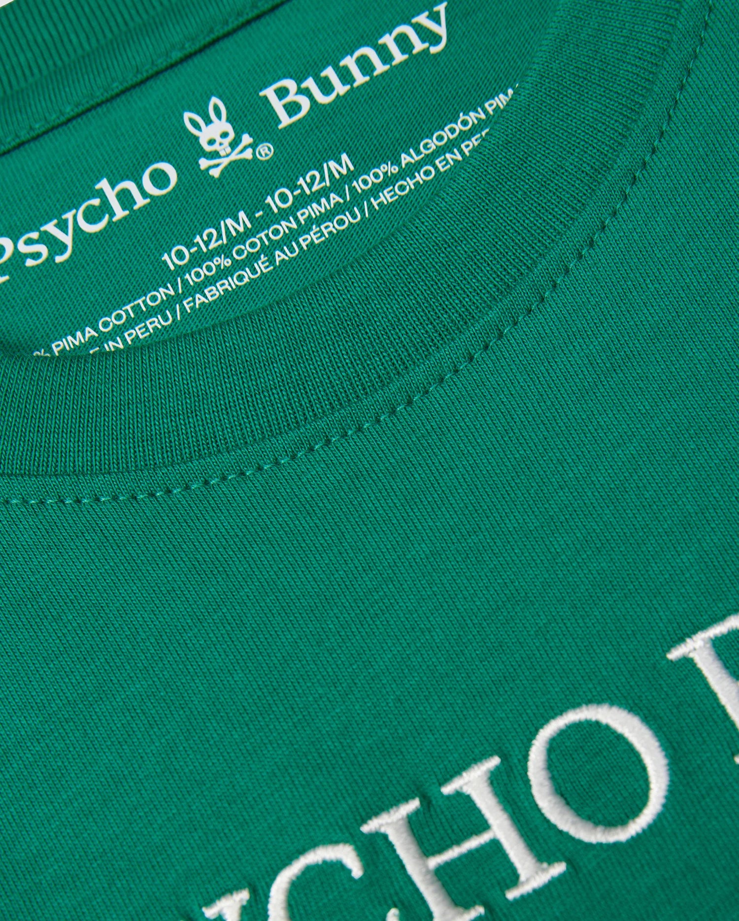 A close-up of a green KIDS COURTSIDE GRAPHIC TEE - B0U684C200 by Psycho Bunny. The text on the shirt's collar reads "Psycho Bunny" with a bunny logo above it. Below it states, "10-12/M, 100% Pima cotton jersey, Made in Peru." The shirt has white embroidered text that is partially visible at the bottom of the image.