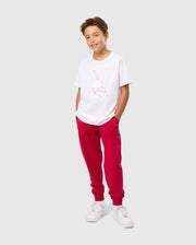 A young boy with short brown hair stands against a plain background, smiling. He wears a white KIDS VINCENT GRAPHIC TEE - B0U713D200 from Psycho Bunny made of soft Pima cotton, featuring a bunny logo. He pairs it with red sweatpants, his hands tucked into his pockets, and completes the timeless look with white sneakers.