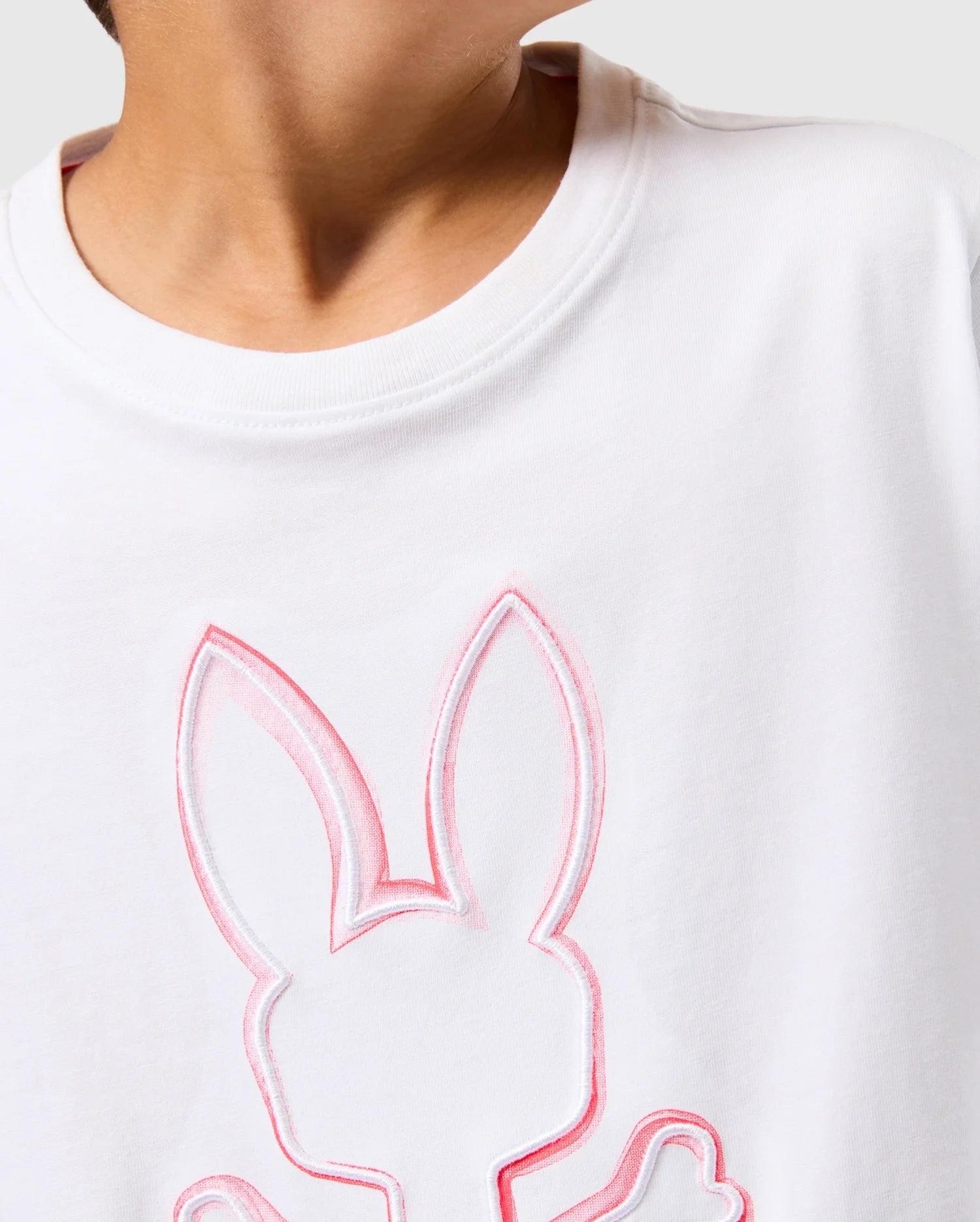 Close-up of a child wearing a white Pima cotton t-shirt with a faint outline of a bunny's head and ears in subtle pink, showcasing the unfadable style of the Psycho Bunny KIDS VINCENT GRAPHIC TEE - B0U713D200. The focus is on the upper torso and the design on the t-shirt against a plain, light grey background.