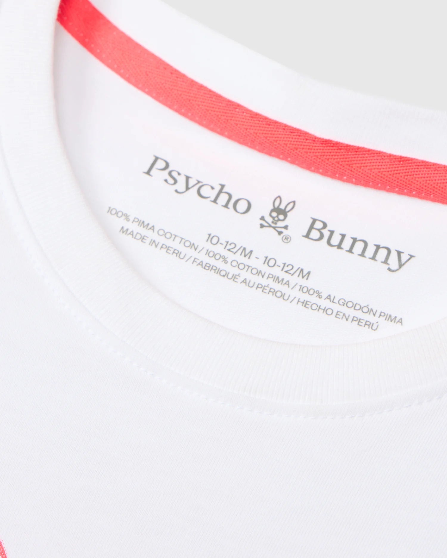 Close-up of a white garment's label, displaying the brand name "Psycho Bunny" along with two stylized bunny ears and skull logo. The label indicates that the KIDS VINCENT GRAPHIC TEE - B0U713D200 is crafted from 100% Pima cotton, sized 10-12/M, and made in Peru with information provided in both English and French.