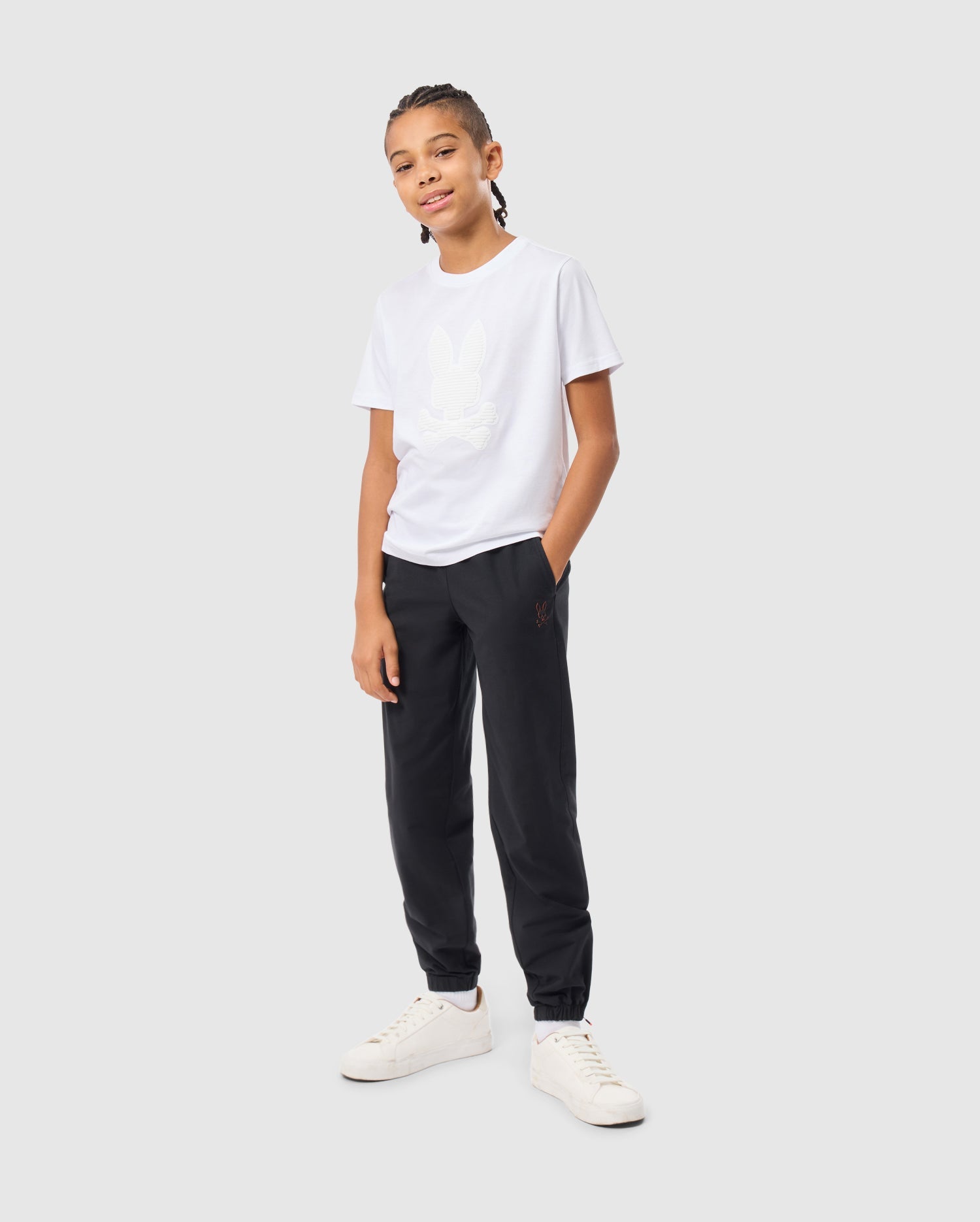 A young individual with braided hair stands confidently, sporting a crisp Psycho Bunny Kids Kingston Graphic Tee crafted from luxurious Pima cotton, paired with dark pants and white sneakers. With one hand in their pocket, they smile at the camera against a plain backdrop.