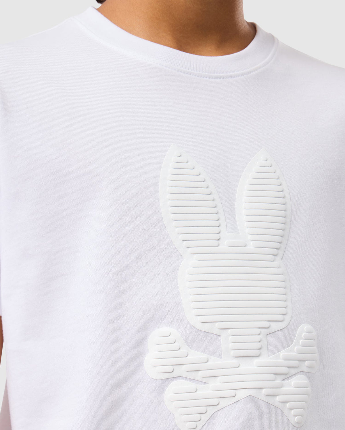 A person is wearing the KIDS KINGSTON GRAPHIC TEE - B0U743D200 by Psycho Bunny, which features an embossed cartoon-style bunny graphic with crossbones. This Pima cotton T-shirt has a round neck and a plain gray background, highlighting only the upper body.
