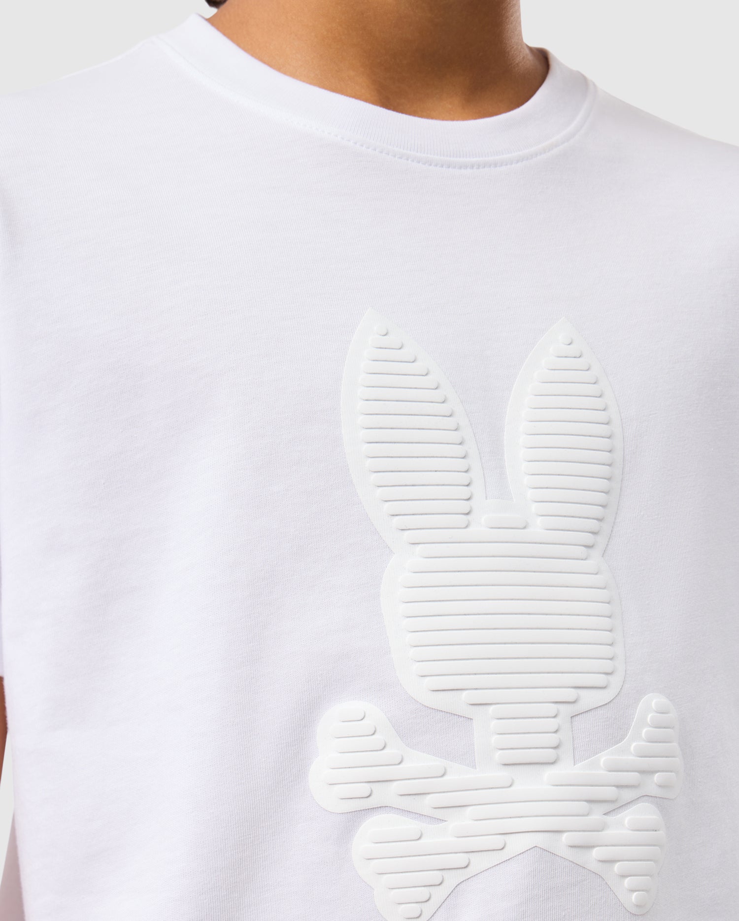 A person is wearing the KIDS KINGSTON GRAPHIC TEE - B0U743D200 by Psycho Bunny, which features an embossed cartoon-style bunny graphic with crossbones. This Pima cotton T-shirt has a round neck and a plain gray background, highlighting only the upper body.